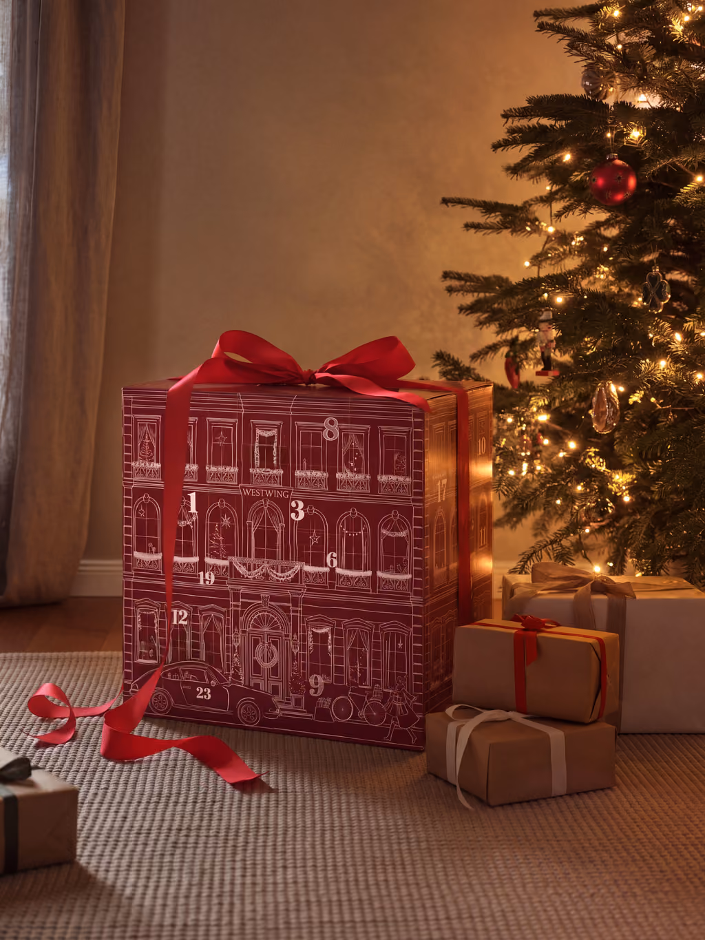 The box tied with a red ribbon on a rug beside a lit Christmas tree and wrapped presents
